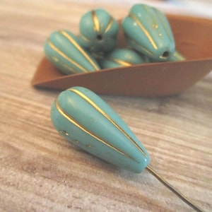 May include: Teardrop-shaped beads in a matte turquoise color featuring vertical gold-colored stripes. One bead is positioned in the foreground on a thin wire, while several additional matching beads are contained within a small brown scoop.