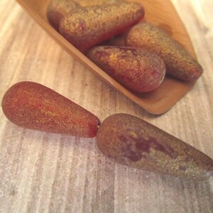 May include: A collection of teardrop-shaped beads with a matte, textured finish in shades of deep red and brown, accented with a metallic gold dusting. Several beads are contained within a small wooden scoop, while two beads are placed outside.