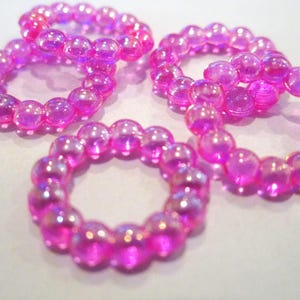 May include: Three circular rings constructed from small, translucent, iridescent pink spherical beads. Each ring features a continuous loop of these shiny, light-reflecting beads, creating a textured, circular shape with a hollow center.