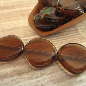 May include: Close-up of brown, translucent glass beads. Three beads are strung together, with a few more in a wooden scoop in the background. The beads are a flat, rounded shape with a hole for stringing.