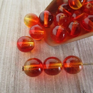 May include: A collection of orange and yellow glass beads. The beads are round with a striped design. Some beads are loose, while three are strung on a gold-colored pin. The beads are displayed on a light-colored wooden surface.