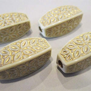 May include: Four off-white oval-shaped beads with a gold floral design. The beads have a hole through the center.