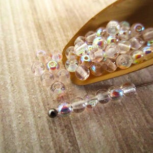 May include: A collection of small, round, clear glass beads with an iridescent finish. Some beads are contained within a wooden scoop, while others are arranged in a single line on a surface, ending with a single dark metallic bead.