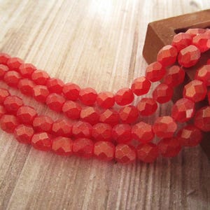 May include: Strands of faceted, red beads. The beads have an oval shape and are strung together. The beads are resting on a light-colored wooden surface. A small, wooden box is in the background.