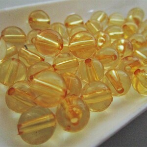 May include: A collection of translucent yellow beads with a shiny finish. The beads are round and have a hole through the center. They are arranged in a pile on a white surface.