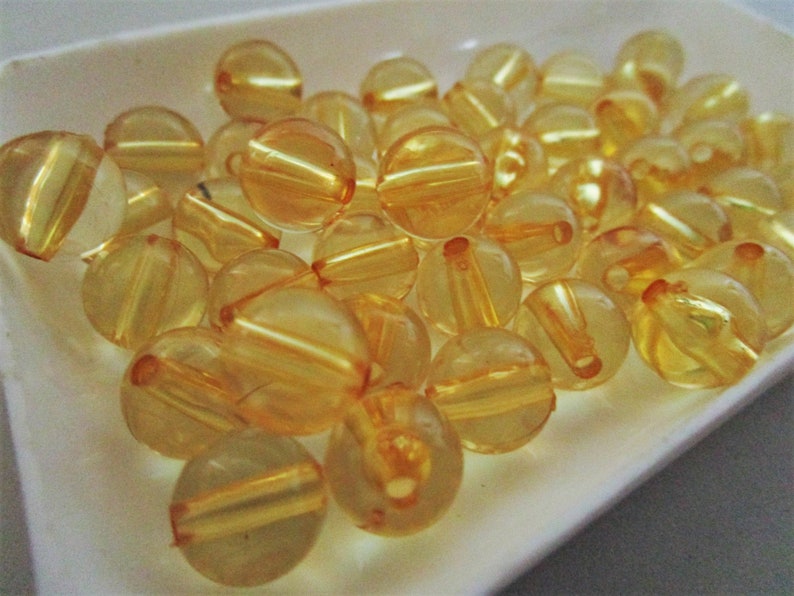 May include: A collection of translucent yellow beads with a shiny finish. The beads are round and have a hole through the center. They are arranged in a pile on a white surface.