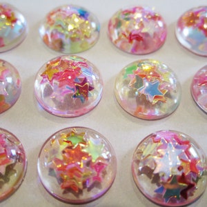 May include: A collection of 12 clear, round, dome-shaped embellishments with iridescent glitter stars inside. The embellishments are pink and have a glossy finish.