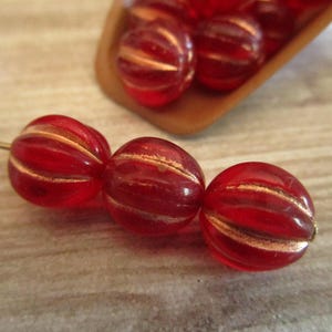 May include: Close-up of three red glass beads with gold stripes, strung on a wire. Additional beads are visible in a wooden scoop in the background. The beads have a ribbed texture.