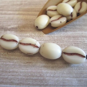 May include: A collection of off-white, oval-shaped beads resembling coffee beans, each featuring a central brown groove. Four of these beads are strung together on a thin metal wire, while several others are contained within a small wooden scoop.