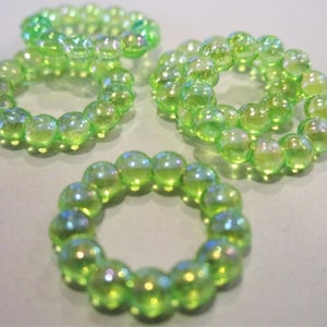 May include: Four rings made of iridescent green glass beads. The beads are round and have a shiny, reflective surface. The rings are arranged on a light gray surface, with the beads catching the light and creating a shimmering effect.