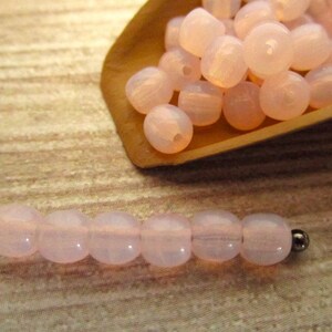 May include: A collection of pale pink, round beads, some strung on a wire, and others in a small wooden scoop. The beads have a translucent quality, and the strung beads are capped with a silver-colored bead.