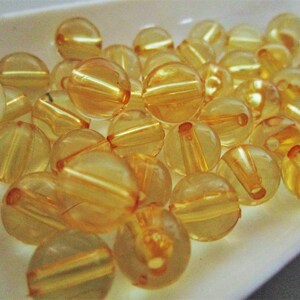 May include: A collection of translucent amber-colored round beads with a shiny finish. The beads have a slightly iridescent quality and are arranged in a pile on a white surface.