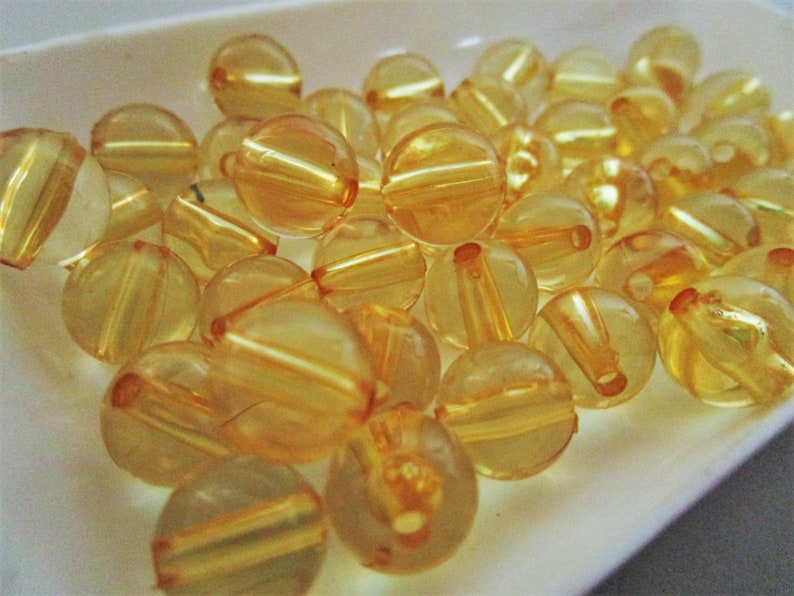 May include: A collection of translucent amber-colored round beads with a shiny finish. The beads have a slightly iridescent quality and are arranged in a pile on a white surface.