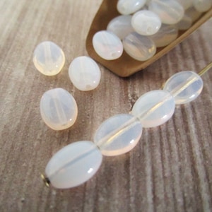 May include: A collection of white oval-shaped glass beads. The beads are strung on a wire and are arranged in a line. The beads are translucent and have a slight iridescent sheen.
