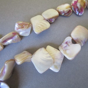 May include: Close-up of a strand of iridescent shell beads. The beads are a creamy white color with hints of pink and purple. The beads are irregularly shaped and have a glossy finish.