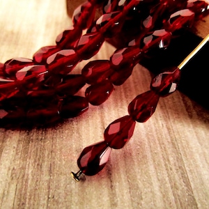 May include: Close-up of a strand of faceted, deep red beads. The beads are teardrop-shaped and strung together. A gold-colored pin holds a few beads, showcasing the design. The beads are on a wooden surface.