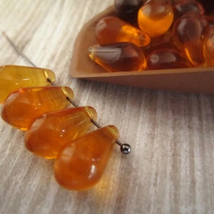 May include: Close-up of amber-colored teardrop-shaped beads strung on a silver wire. A small wooden scoop holds more of the same beads. The beads have a translucent quality and are arranged on a light-colored, textured surface.