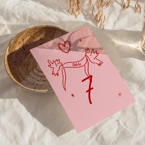 May include: A pink table number card with red illustrations of two birds holding a banner that reads "table" and the number "7". A heart and stars are also drawn on the card. The card is on a white surface.