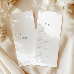 Puede incluir: Dos tarjetas de menú de boda blancas con texto negro. Una tarjeta dice "Katie and Joseph" y la otra "Menú" con una lista de alimentos. Las tarjetas están sobre una tela color crema con flores blancas.