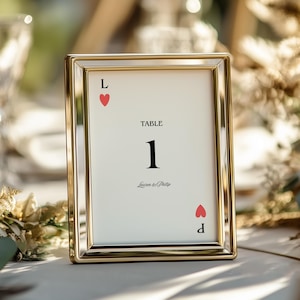 May include: A gold framed table number card with the text "TABLE 1" in black. The card has a white background with a red heart and the letters "L" and "d" on the top and bottom. The names "Lauren & Phillip" are at the bottom.