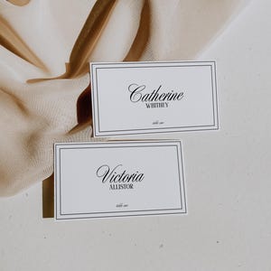 May include: Two white place cards with black borders and black cursive text. The top card reads "Catherine Whitney, table one". The bottom card reads "Victoria Allister, table one".