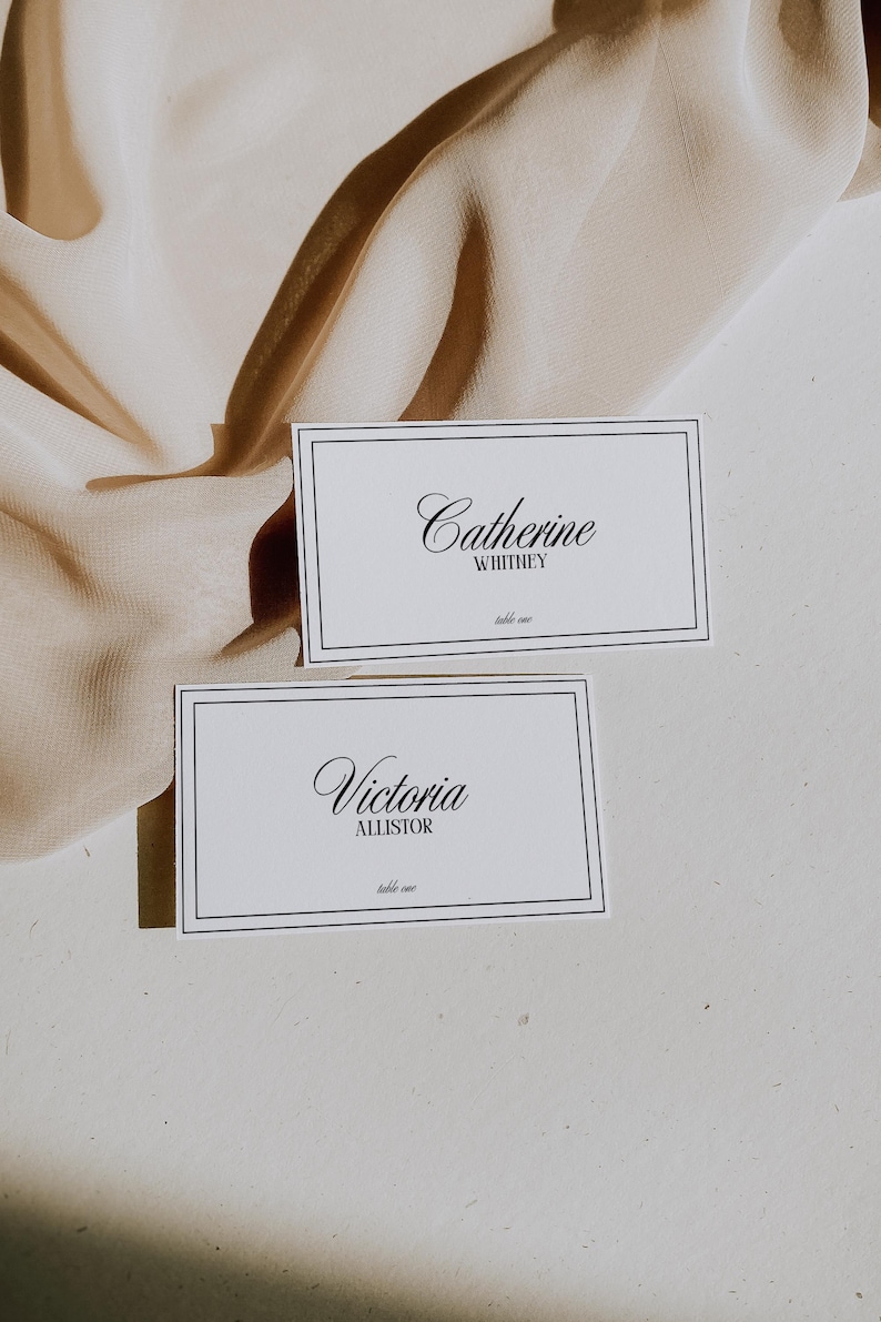 May include: Two white place cards with black borders and black cursive text. The top card reads "Catherine Whitney, table one". The bottom card reads "Victoria Allister, table one".