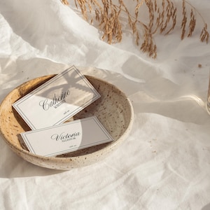 May include: Two white business cards with black text in a small, speckled, beige bowl. The top card reads "Catherine Whittier" and the bottom card reads "Victoria Alexander".