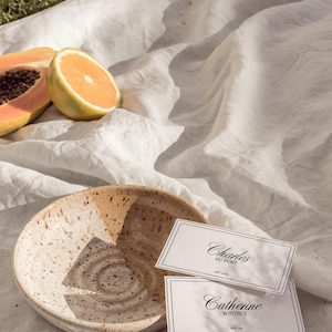 May include: Two white place cards with black calligraphy on a white linen tablecloth. The top card reads "Charles Du Pont" and the bottom card reads "Catherine Whitney". A brown and white ceramic bowl and pieces of papaya and orange are in the background.
