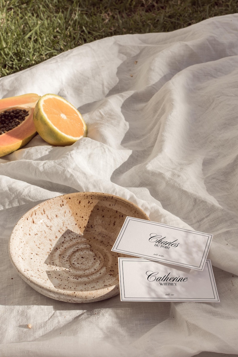 May include: Two white place cards with black calligraphy on a white linen tablecloth. The top card reads "Charles Du Pont" and the bottom card reads "Catherine Whitney". A brown and white ceramic bowl and pieces of papaya and orange are in the background.