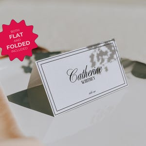 May include: White place card with a black border and text that reads "Catherine Whitney" in a cursive font. The place card is sitting on a white surface with a shadow underneath.