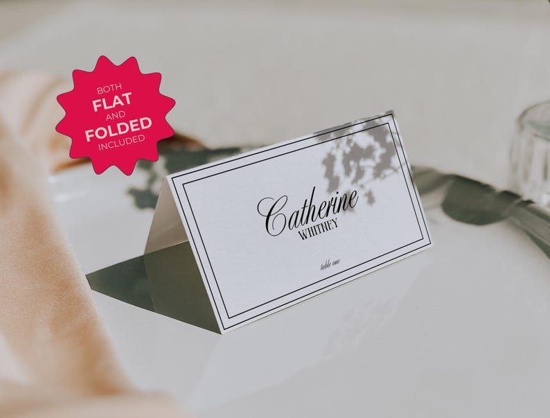 May include: White place card with a black border and text that reads "Catherine Whitney" in a cursive font. The place card is sitting on a white surface with a shadow underneath.