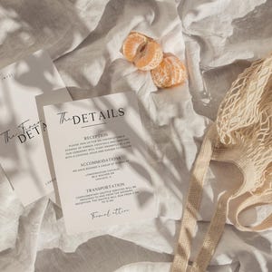 May include: Two white cards with black text on a white linen background. The text on the cards reads "The First Details" and "The Details". The card on the right has a list of details including "Reception", "Accommodation", "Transportation", and "Final Location".