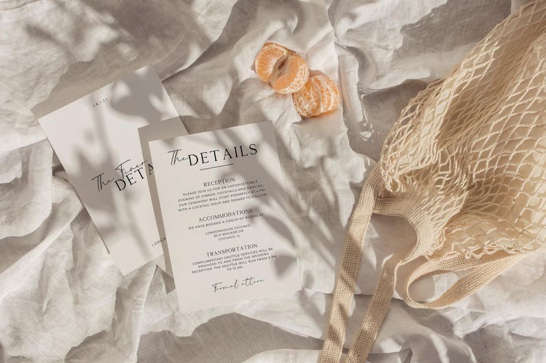 May include: Two white cards with black text on a white linen background. The text on the cards reads "The First Details" and "The Details". The card on the right has a list of details including "Reception", "Accommodation", "Transportation", and "Final Location".