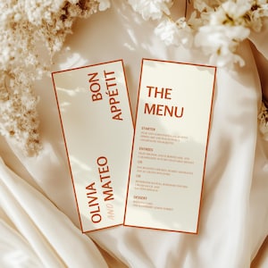 Może przedstawiać: Dwie kremowe karty menu weselnego z jasnobrązową obwódką. Na jednej karcie widnieje napis "BON APPÉTIT" i "OLIVIA AND MATEO". Na drugiej karcie widnieje napis "THE MENU" z listą pozycji menu. Karty leżą na białej satynowej powierzchni.