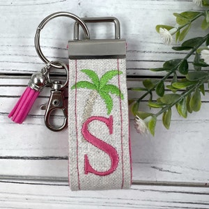 May include: A white and pink fabric keychain with a silver key ring and a pink tassel. The fabric has an embroidered palm tree and the letter "S".