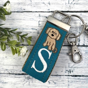 May include: Teal blue fabric keychain with a white embroidered letter "S" and a brown embroidered dog. The keychain has a silver metal ring and clasp.