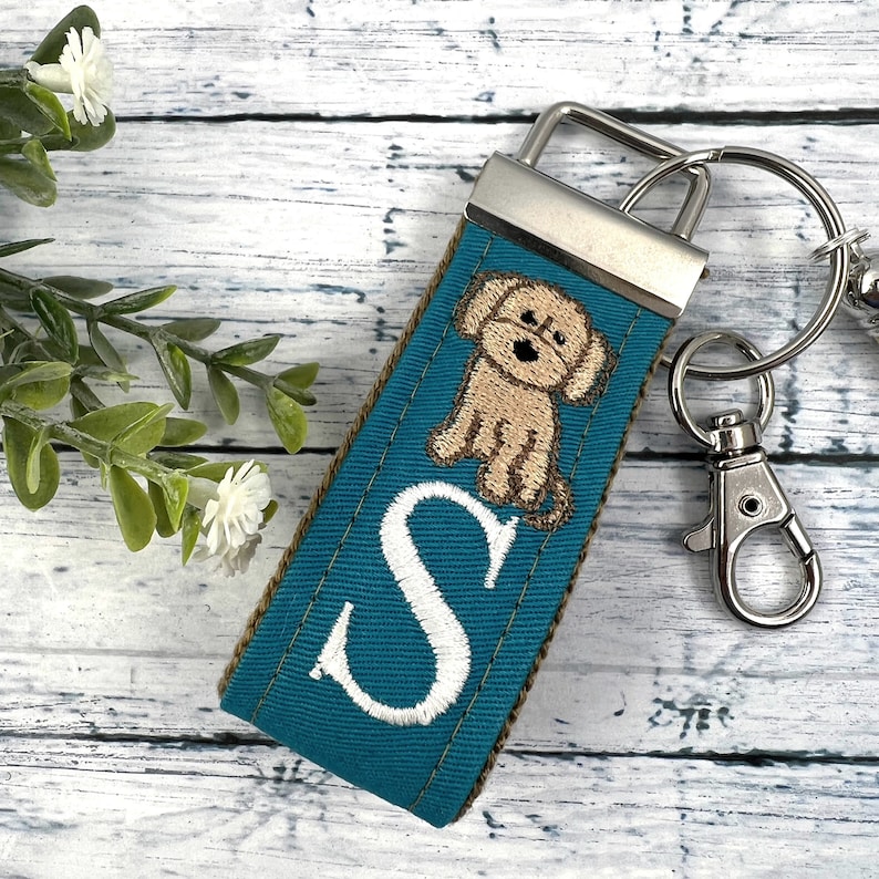 May include: Teal blue fabric keychain with a white embroidered letter "S" and a brown embroidered dog. The keychain has a silver metal ring and clasp.