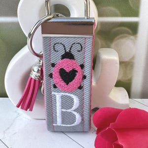 May include: A gray and pink keychain with a ladybug design and the letter "B". The ladybug has a pink body with a black heart and black spots. A pink tassel hangs from the silver-colored metal hardware.