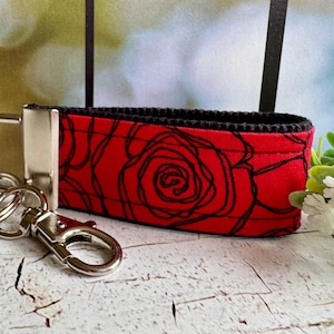 May include: A red fabric keychain wristlet with a black rose outline design. The wristlet has a black border and a silver-colored metal clasp and key ring. The background is blurred, with a hint of greenery.