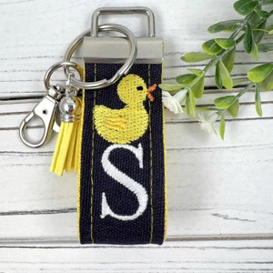 May include: A black and yellow key chain with a yellow rubber ducky and the letter "S" embroidered on it. The key chain has a silver metal ring and a yellow tassel.
