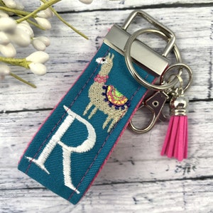 May include: Teal blue fabric keychain with a white embroidered letter R and a beige llama with colorful accents. The keychain has a silver key ring and a pink tassel.