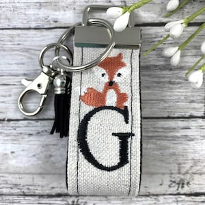 May include: A white fabric keychain with a black embroidered letter G and a small orange fox design. The keychain has a silver metal key ring and a black tassel.