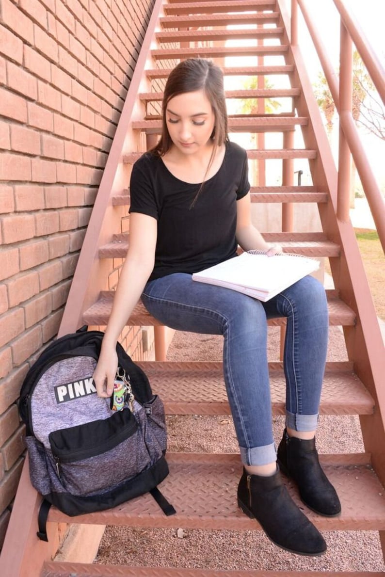 May include: A young person sitting on a set of stairs wearing a black shirt, blue jeans, and black ankle boots. They are holding a notebook and a backpack with the word "PINK" on it.