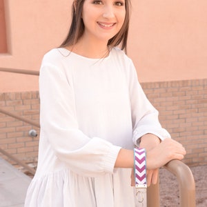 Pu&ograve; includere: Una donna indossa una blusa bianca con volant e jeans blu e tiene un portachiavi con motivo chevron viola e blu con chiavi dorate attaccate.