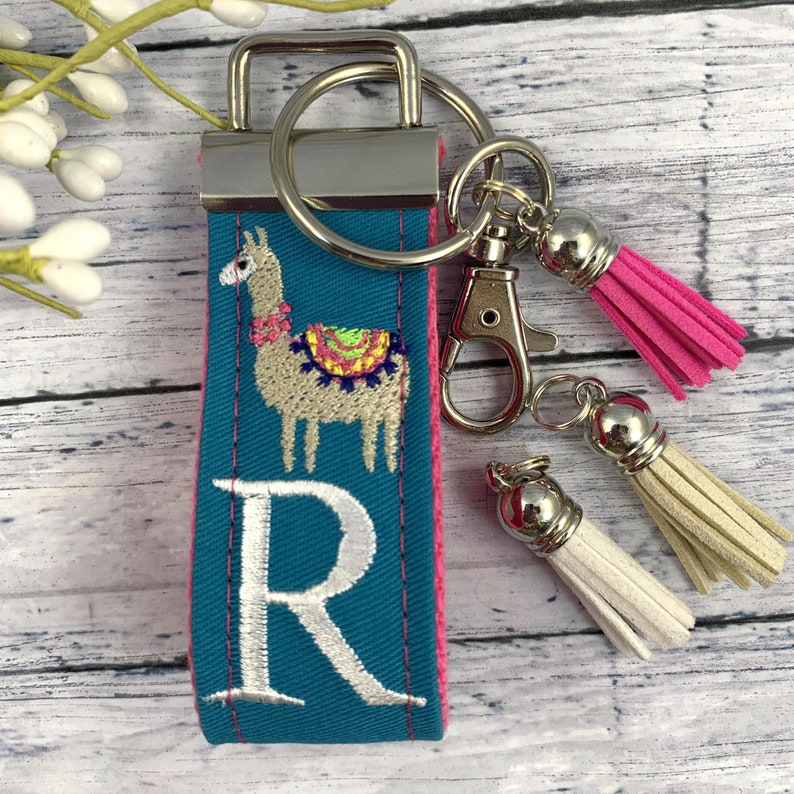May include: Teal blue key chain with a white embroidered letter R and a llama with a colorful saddle. The key chain has a silver key ring and two tassels, one pink and one white.