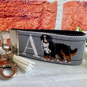 May include: Gray fabric keychain with a silver metal clasp and a white tassel. The keychain features an embroidered Bernese Mountain Dog and the letter "A". The dog is black, brown, and white.