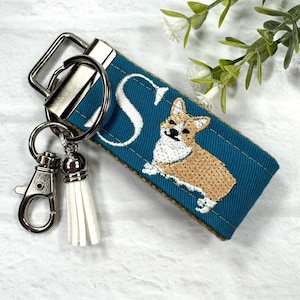 May include: Teal blue fabric key chain with a white embroidered letter "S" and a brown and white embroidered corgi dog. The key chain has a silver metal key ring and a white tassel.