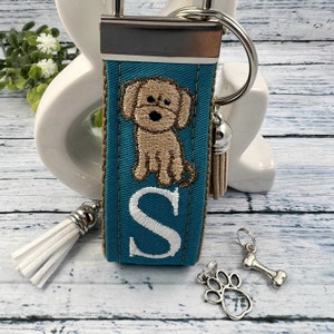 May include: Teal blue fabric keychain with a white letter S, a brown dog embroidery, and a white tassel. The keychain also includes a silver bone charm and a silver paw print charm.