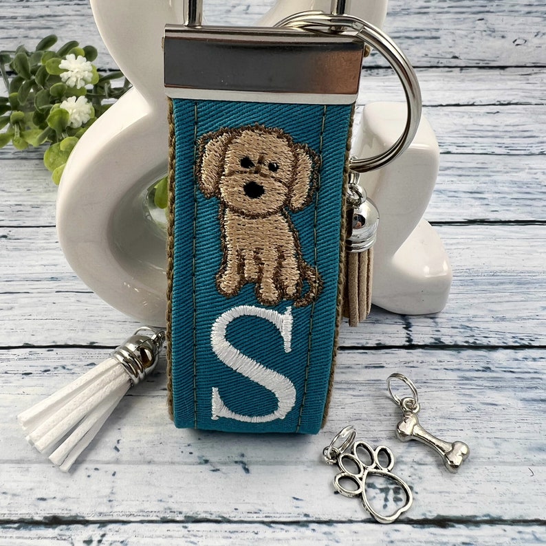 May include: Teal blue fabric keychain with a white letter S, a brown dog embroidery, and a white tassel. The keychain also includes a silver bone charm and a silver paw print charm.