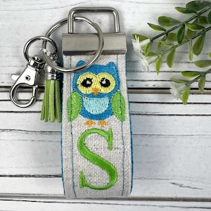 May include: A white fabric keychain with a green and blue embroidered owl and the letter 'S' in green. The keychain has a silver key ring and a green tassel.